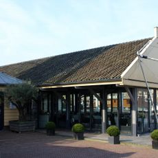 Langgerekte, open loods, opgetrokken in hout - met staand beschot - onder flauwhellend zadeldak met groot overstek tussen puntgevels, met uitzwenking naar de top.