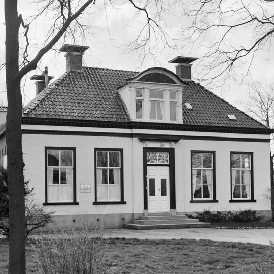 Landelijk huis met omlijste ingang en zesruitsvensters onder omgaand schilddak met versierde dakkapel en vier hoekschoorstenen met borden