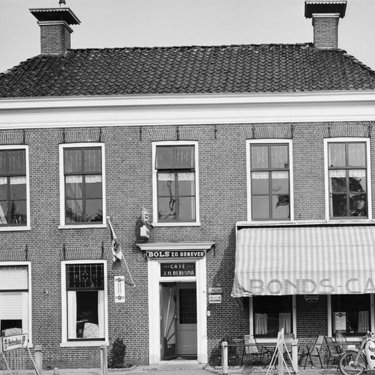 Blokvormig pand met omlijste ingang en verdieping met zesruitsvensters onder laag schilddak met twee hoekschoorstenen met borden