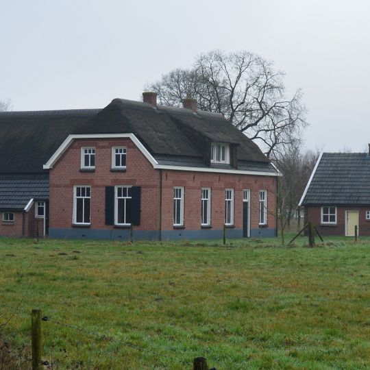 Boerderij op een T-vormige plattegrond opgetrokken in rode baksteen, onder een deels met riet, deels met grijze muldenpannen gedekt wolfdak met rieten dakruiters