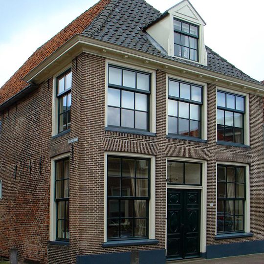 Ten dele onderkelderd hoekpand met verdieping en rechte kroonlijst onder schilddak met voordeur en dakkapel in Lodewijk XVI-/Empirestijl