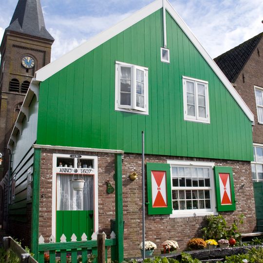 Verhoogd huis onder een met pannen gedekt zadeldak met een eenvoudige topgevel bestaande uit een bakstenen pui en houten top, en houten zijgevels op bakstenen voeting
