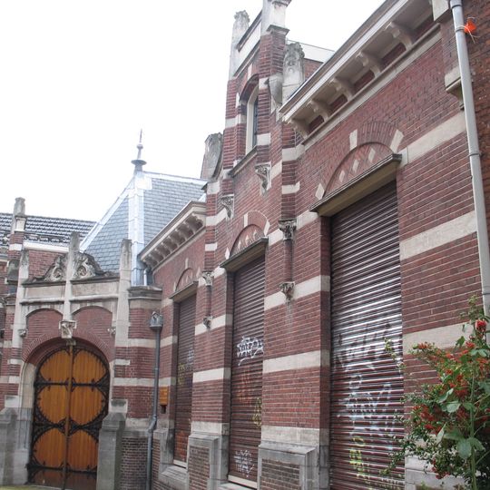 Schuin in de rooilijn staand pand, waarin gehuisvest de Herenkamer, onder afgeplat schilddak met leien gedekt, opgetrokken in baksteen, afgewisseld met natuurstenen blokken en banden.