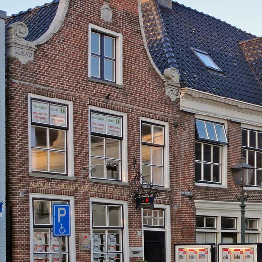 Pand met verdieping onder zadeldak tegen klokgevel in de plaatselijke trant versierd met gebeeldhouwde aanzetkrullen, lijst en kuifstuk