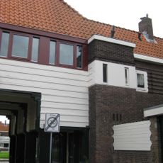 Poortgebouw met twee winkels met bovenwoningen, met veel hout zoals de zware kolommen in poort, bouwstijl Amsterdamse School, ligging aan Purmerweg centraal deel van Tuindorp Nieuwendam