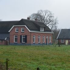Boerderij op een T-vormige plattegrond opgetrokken in rode baksteen, onder een deels met riet, deels met grijze muldenpannen gedekt wolfdak met rieten dakruiters