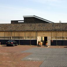 Langgerekte, gesloten loods opgetrokken in hout delen (rabat) onder pannen zadeldak tussen puntgevels. Te linkerzijde aangekapt een aanbouw onder lessenaarsdak