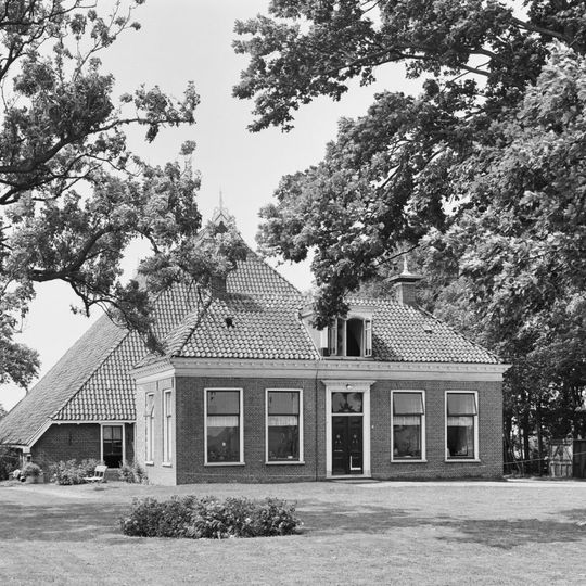 Boerderij met groot schuurdak en dwars voorhuis met omlijste ingang en geblokte kroonlijst onder schilddak met tweedelige dakkapel en twee hoekschoorstenen met versierde borden