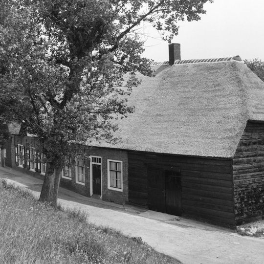 Langwerpige hoeve aan de voet van de dijk met woongedeelte, waarin vensters met negenruitsschuiframen, tussen stallen, waarvan de wanden bestaan uit geteerde houten delen. Strodaken
