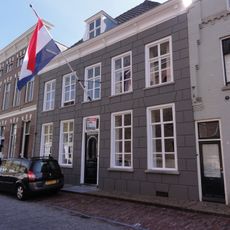 Huis met grijze gebosseerd gepleisterde lijstgevel, kroonlijst op consoles, dwars schilddak, dakkapel en schuiframen, deur