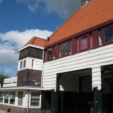 Poortgebouw met twee winkels met bovenwoningen, met veel hout zoals de zware kolommen in poort, bouwstijl Amsterdamse School, ligging aan Purmerweg centraal deel van Tuindorp Nieuwendam