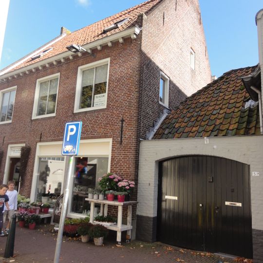 Eenvoudig dorpshuis met verdieping, grijs gecement, beneden met vensterluiken, hoog dwars zadeldak tussen zijtopgevels met vlechtingen