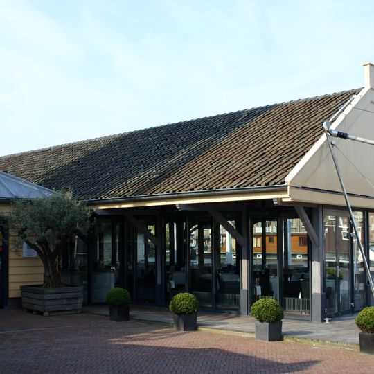 Langgerekte, open loods, opgetrokken in hout - met staand beschot - onder flauwhellend zadeldak met groot overstek tussen puntgevels, met uitzwenking naar de top.